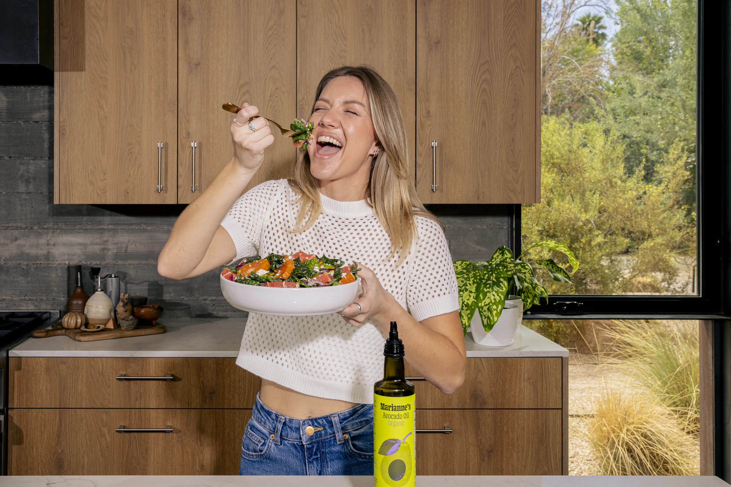 A woman laughs mid-bite over a bowl of citrus salad, the avocado oil bottle in frame beside her