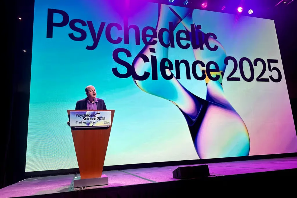 Governor Jared Polis at the podium in front of the keynote screen, the Psychedelic Science 2025 logo refracted through a cyan molecule, Colorado Convention Center, June 18, 2025. Photo: Kiare DeMare, CPR News