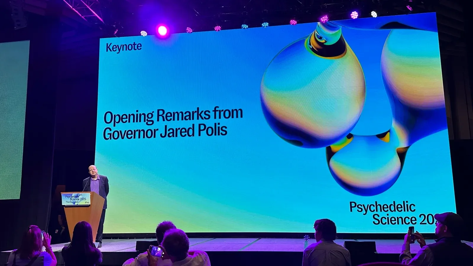 Governor Jared Polis opening Psychedelic Science 2025 under the keynote wall, the molecule on its gradient
